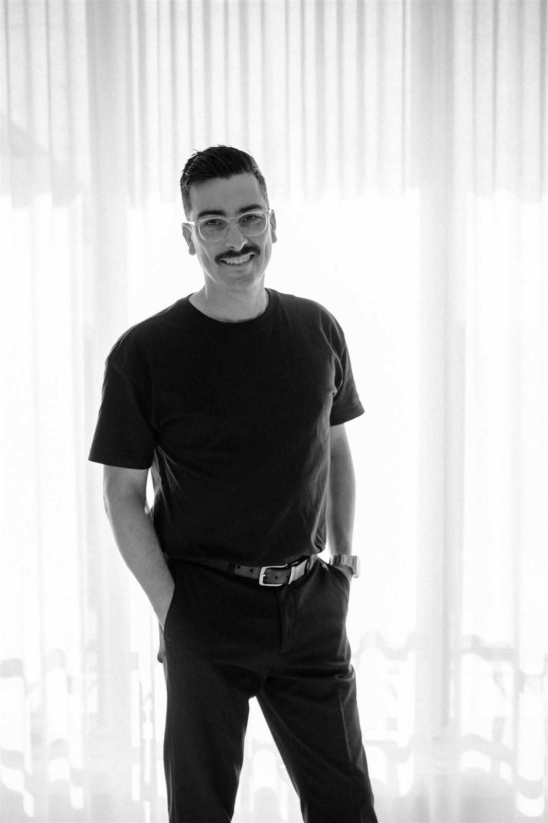 Black and white portrait of Alex Olguin standing in front of sheer curtains