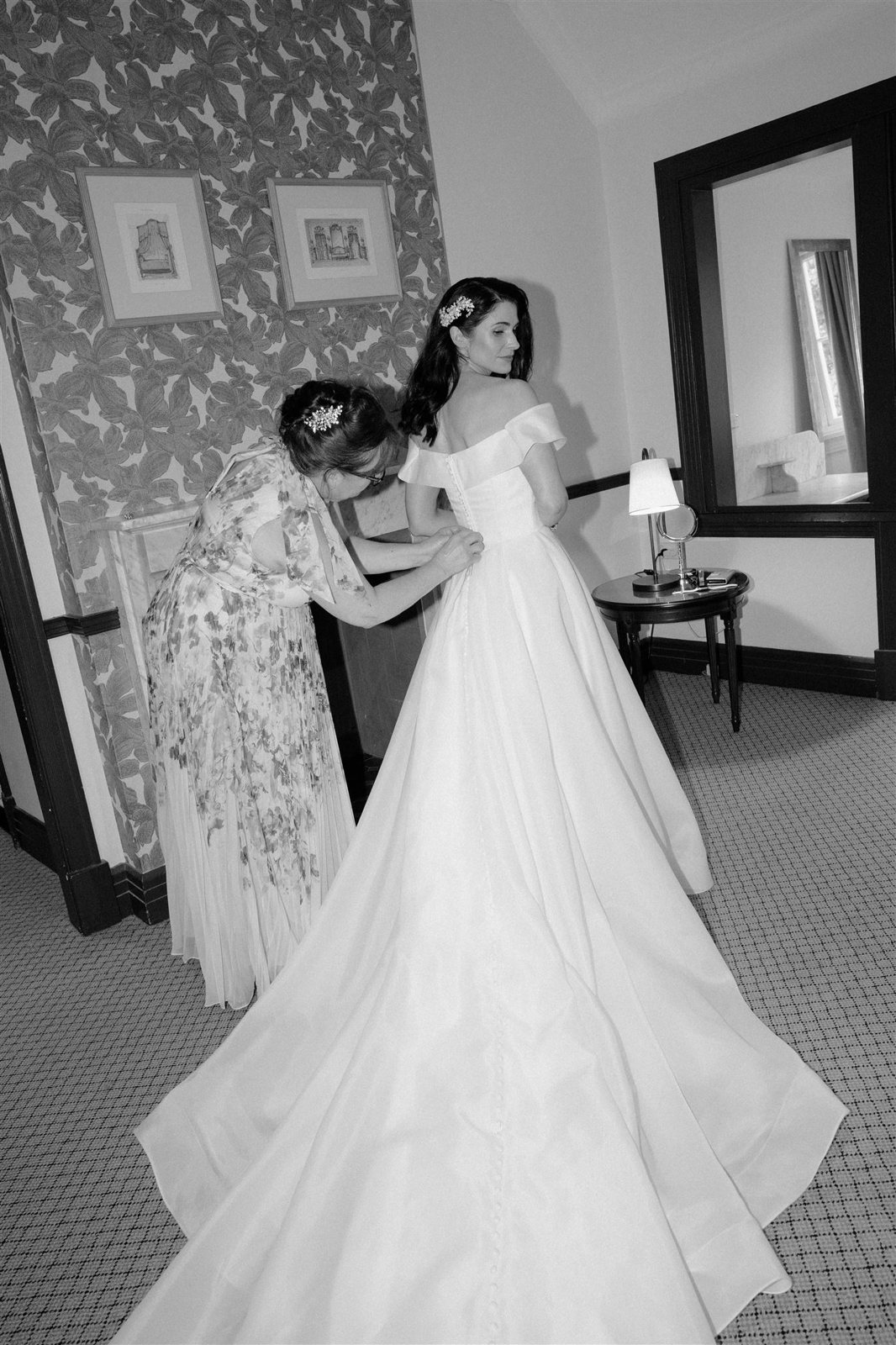 Black and white: a woman fastening the back of a bride&rsquo;s gown in a wallpapered room
