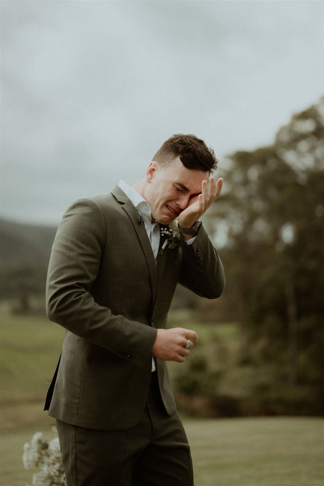 A groom in a green suit wiping away a tear on a lawn