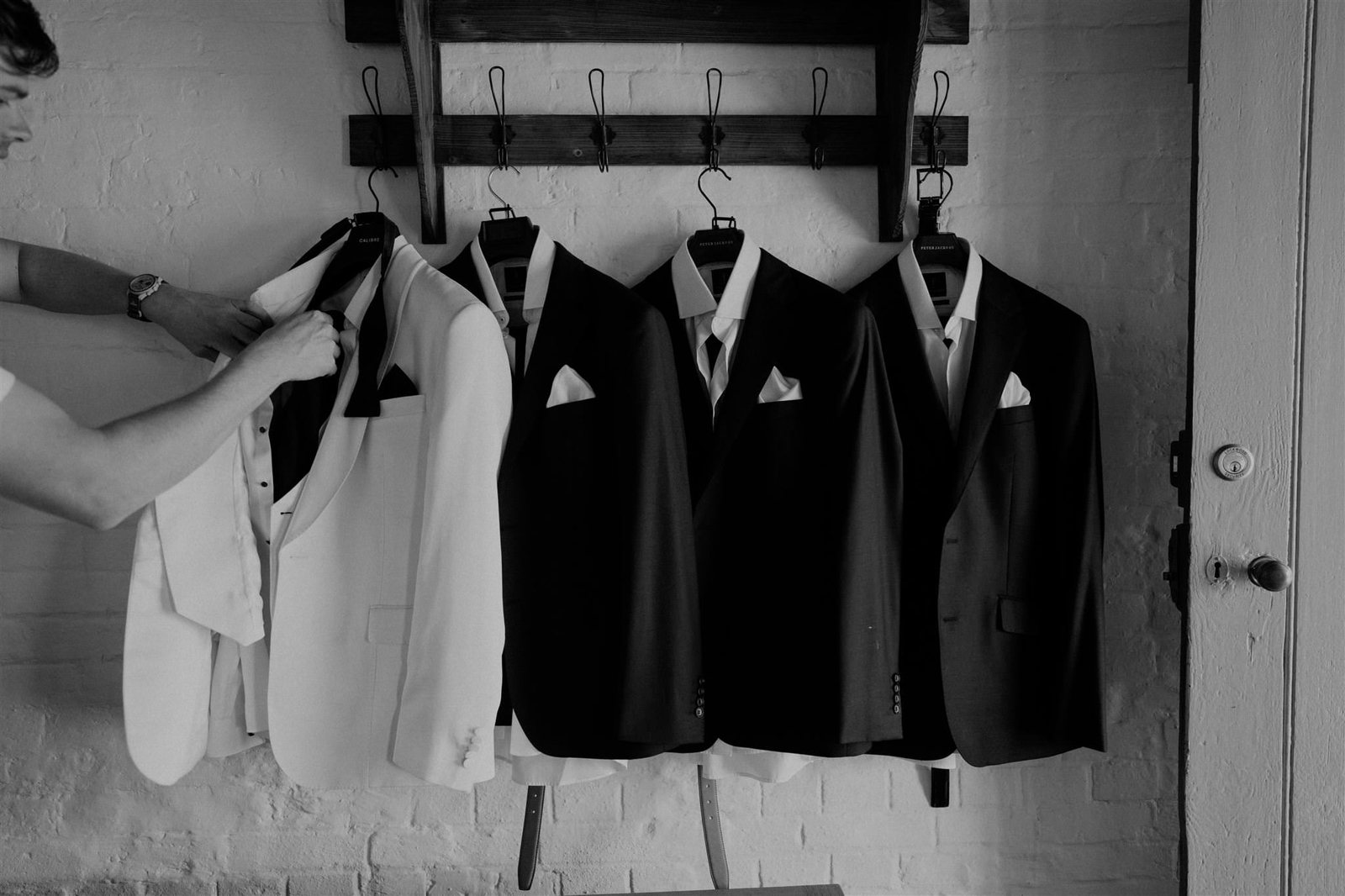 Black and white: suits and a white dinner jacket hanging in a row as a hand straightens a bow tie