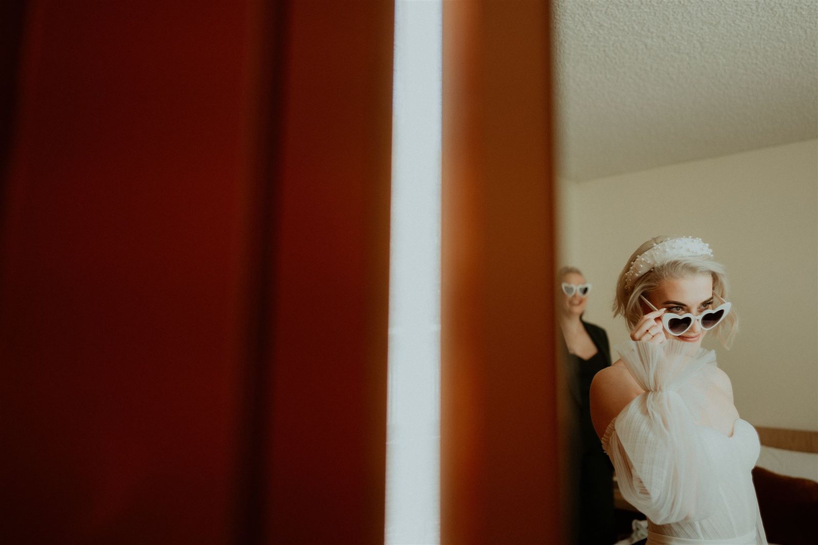 A bride in white sunglasses and a veil framed by a red curtain