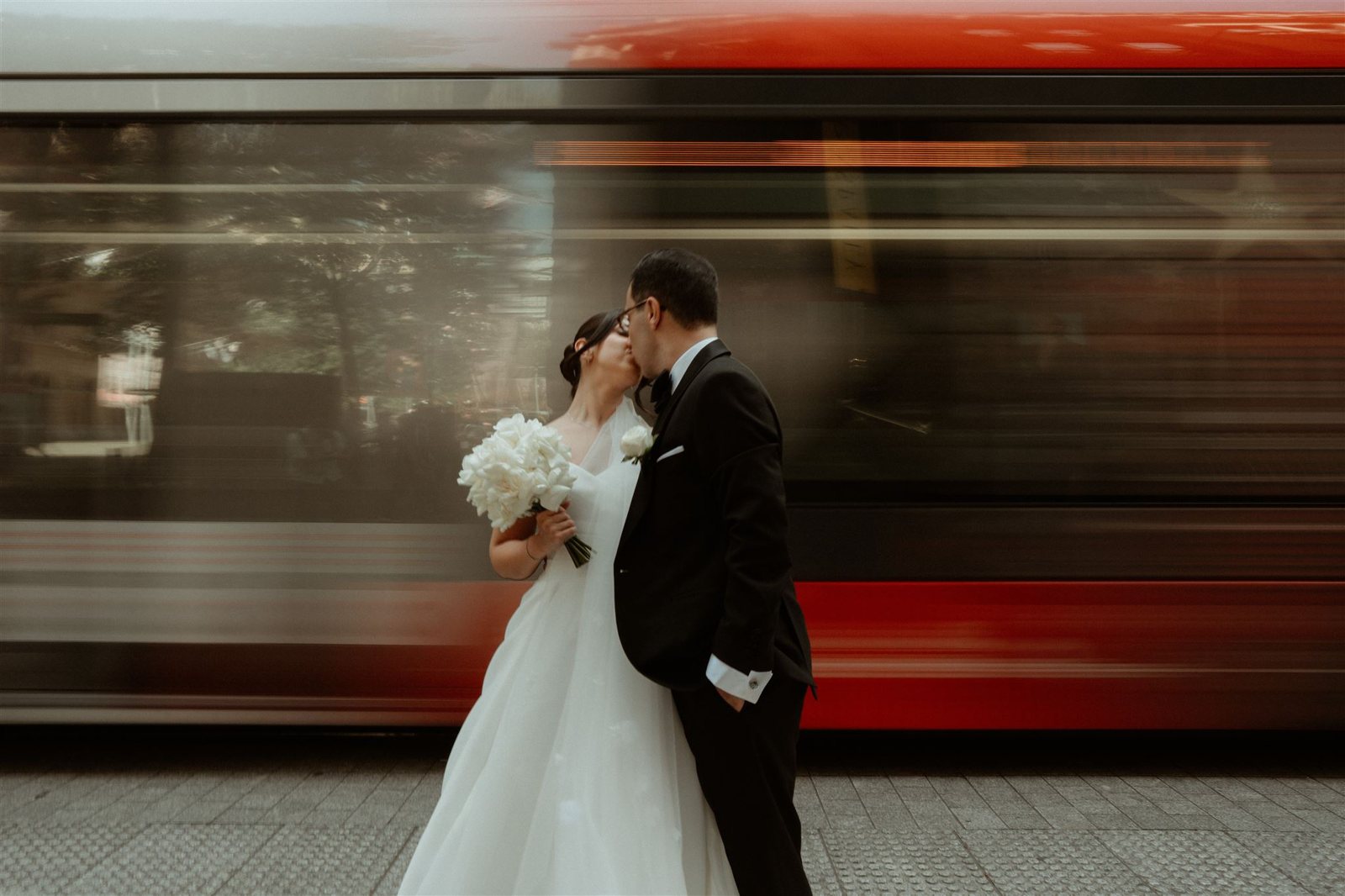 A couple kissing on a station platform as a red train streaks past