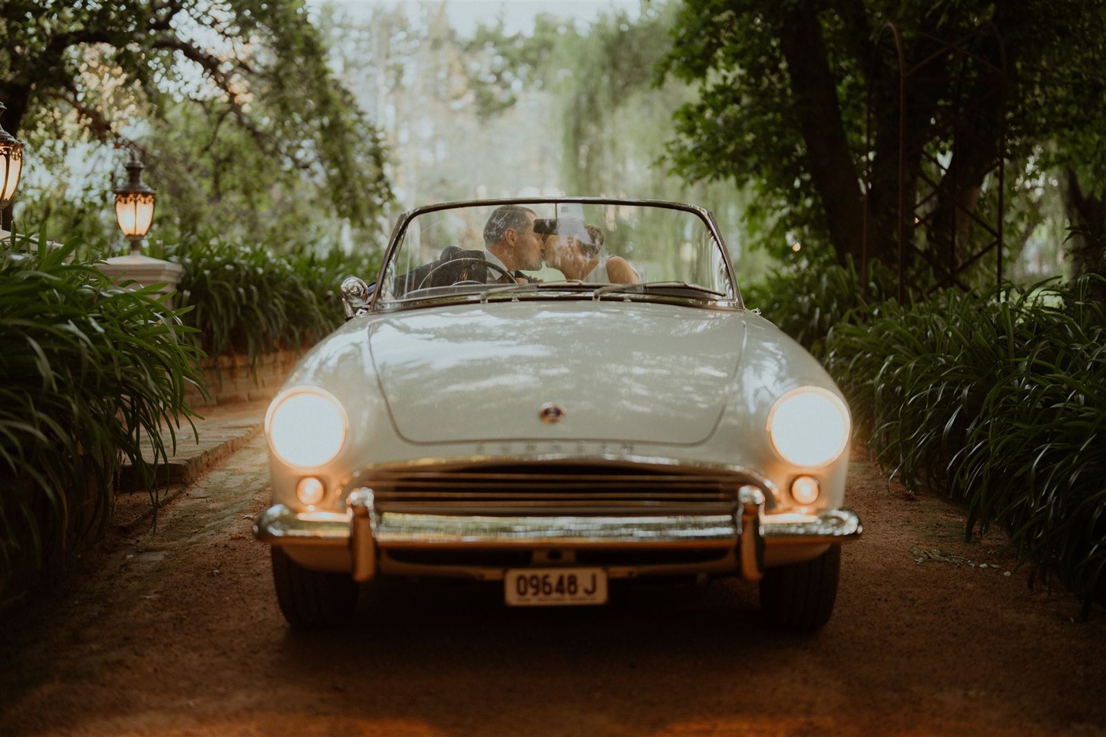 A couple in a white vintage convertible on a leafy driveway